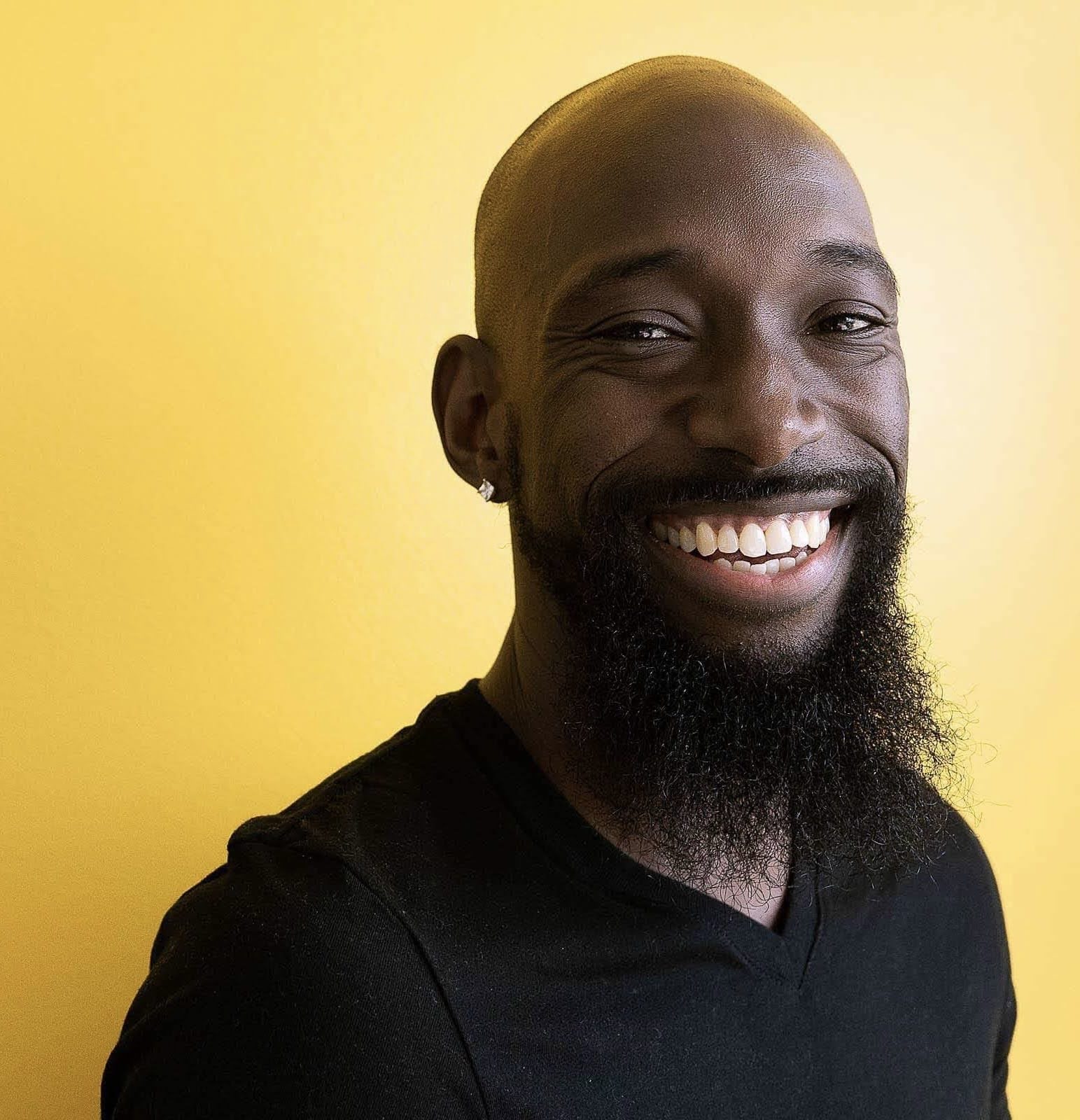 Smiling man with a beard and earring against a yellow background.