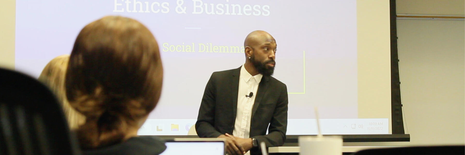Man speaking in front of a presentation screen during a seminar or conference.