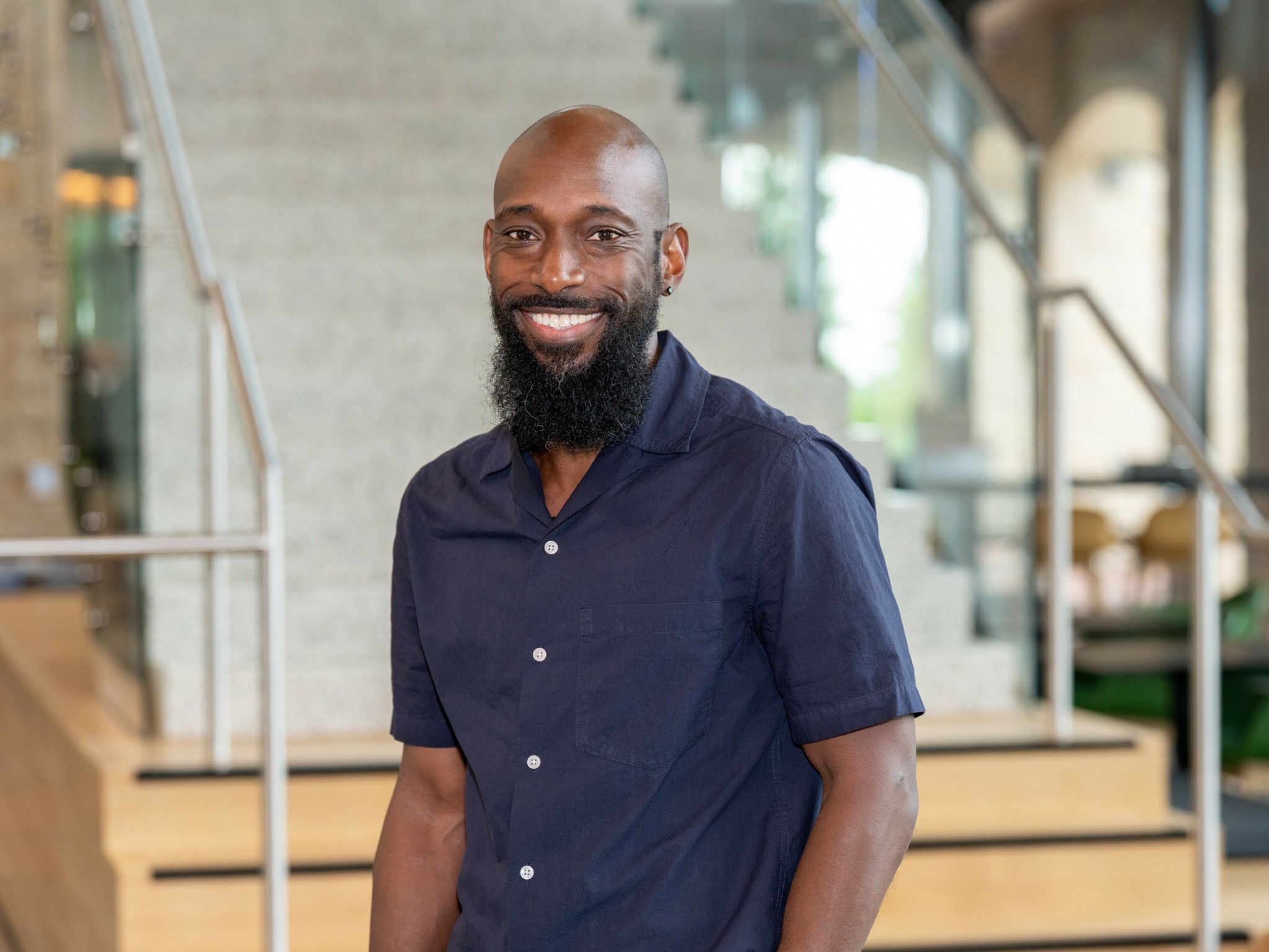 Smiling man with a beard wearing a navy blue short-sleeved shirt indoors.