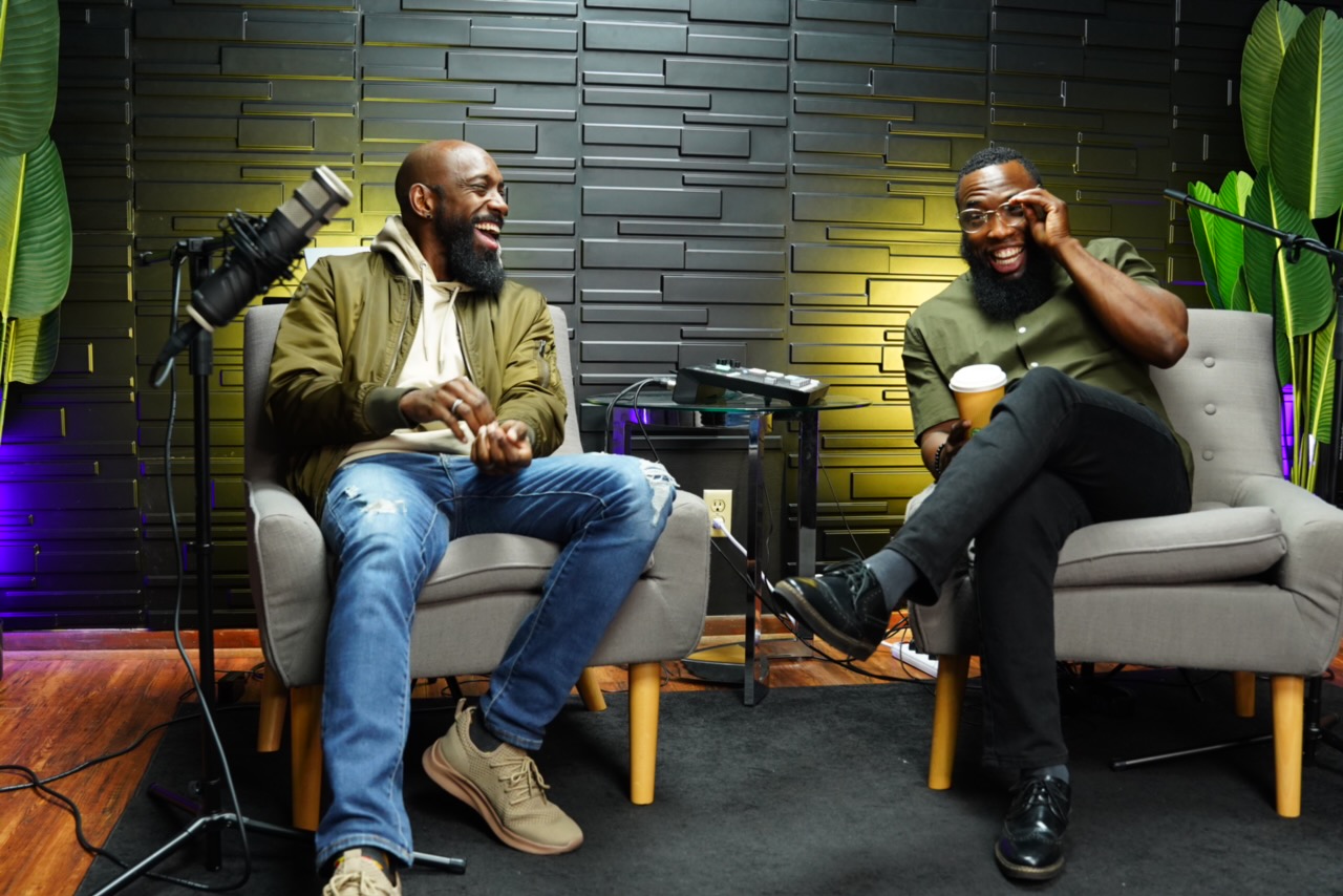 Two men sit on chairs in a podcast studio, smiling and talking, with a microphone and plants in the background.