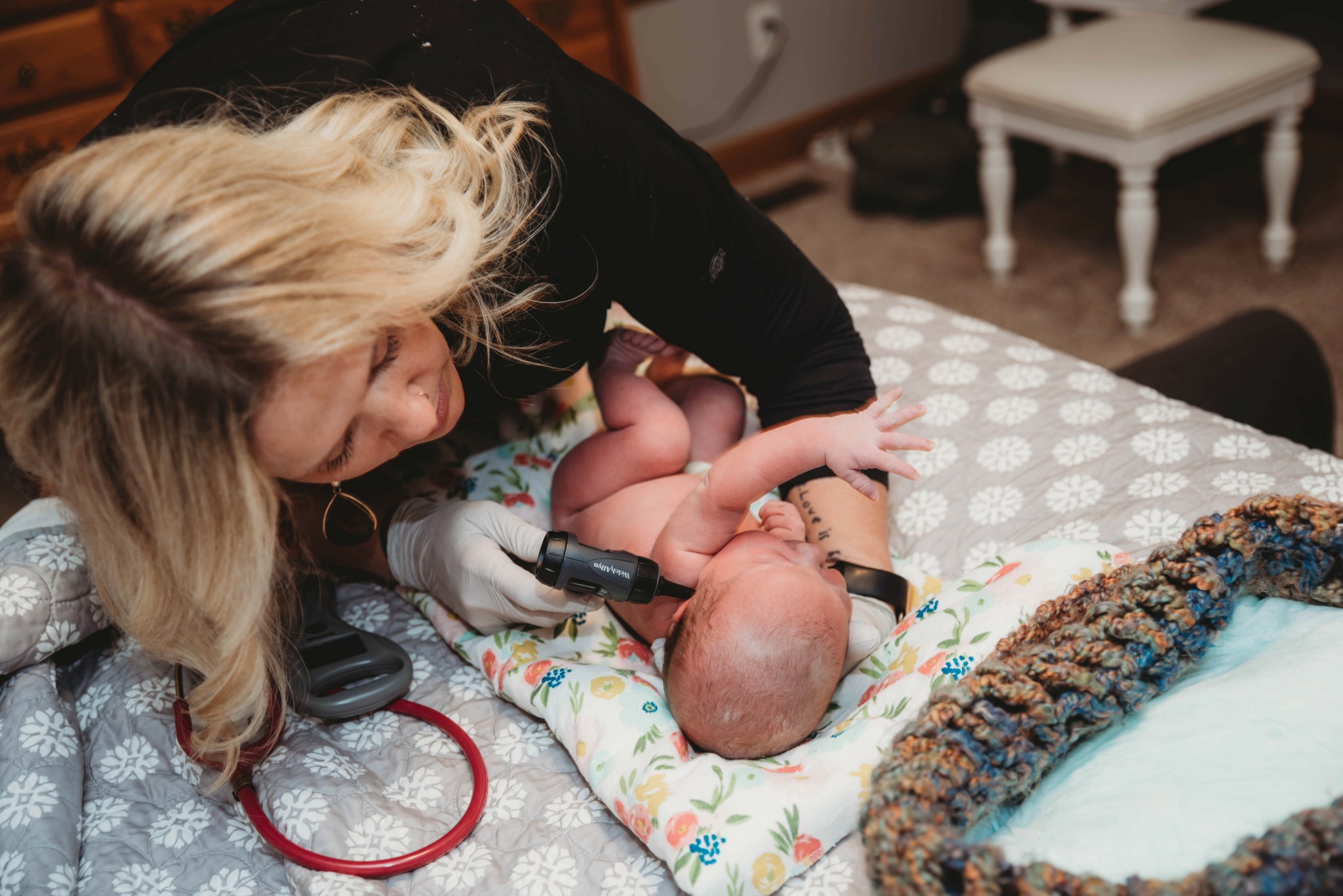 Adult leaning over a baby lying on a patterned blanket on a bed, with a stethoscope nearby and a baby bath in the foreground.