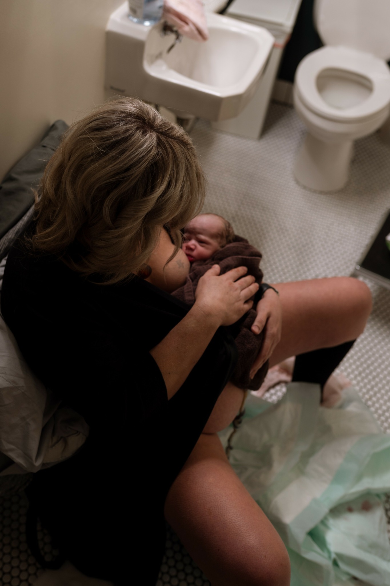 Person sitting on a toilet with a baby in their lap in a bathroom with a sink and toilet.