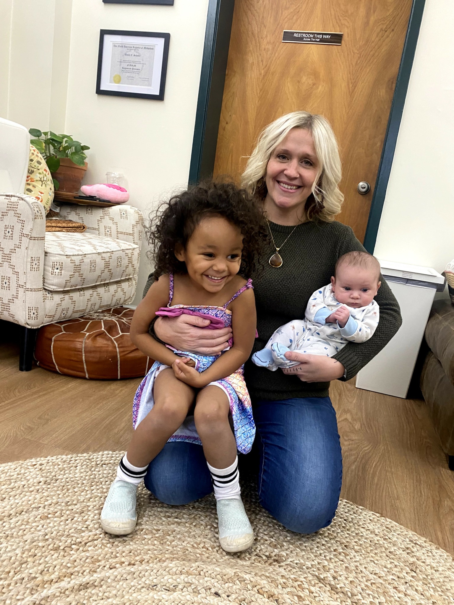 Woman kneeling with a young girl and a baby in a room with a door, framed certificate, and plants.