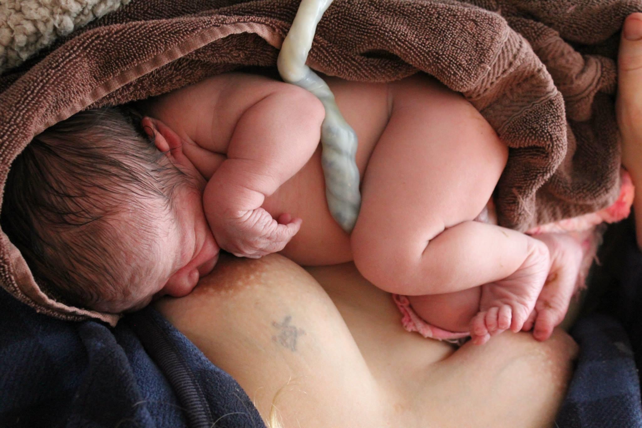 Newborn baby lying on a person's chest, wrapped in a brown blanket, with a medical cord attached.