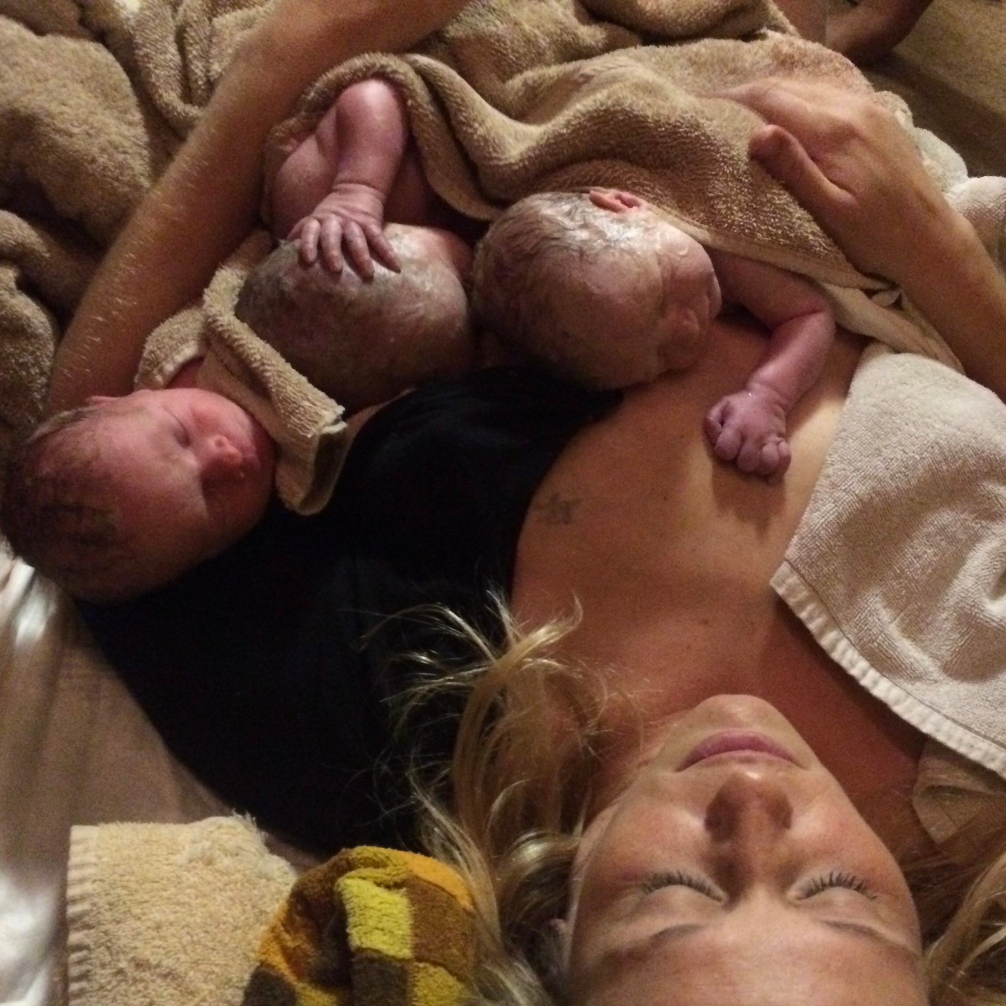 Woman lying down with three newborn babies on her chest, wrapped in towels, in a cozy setting.