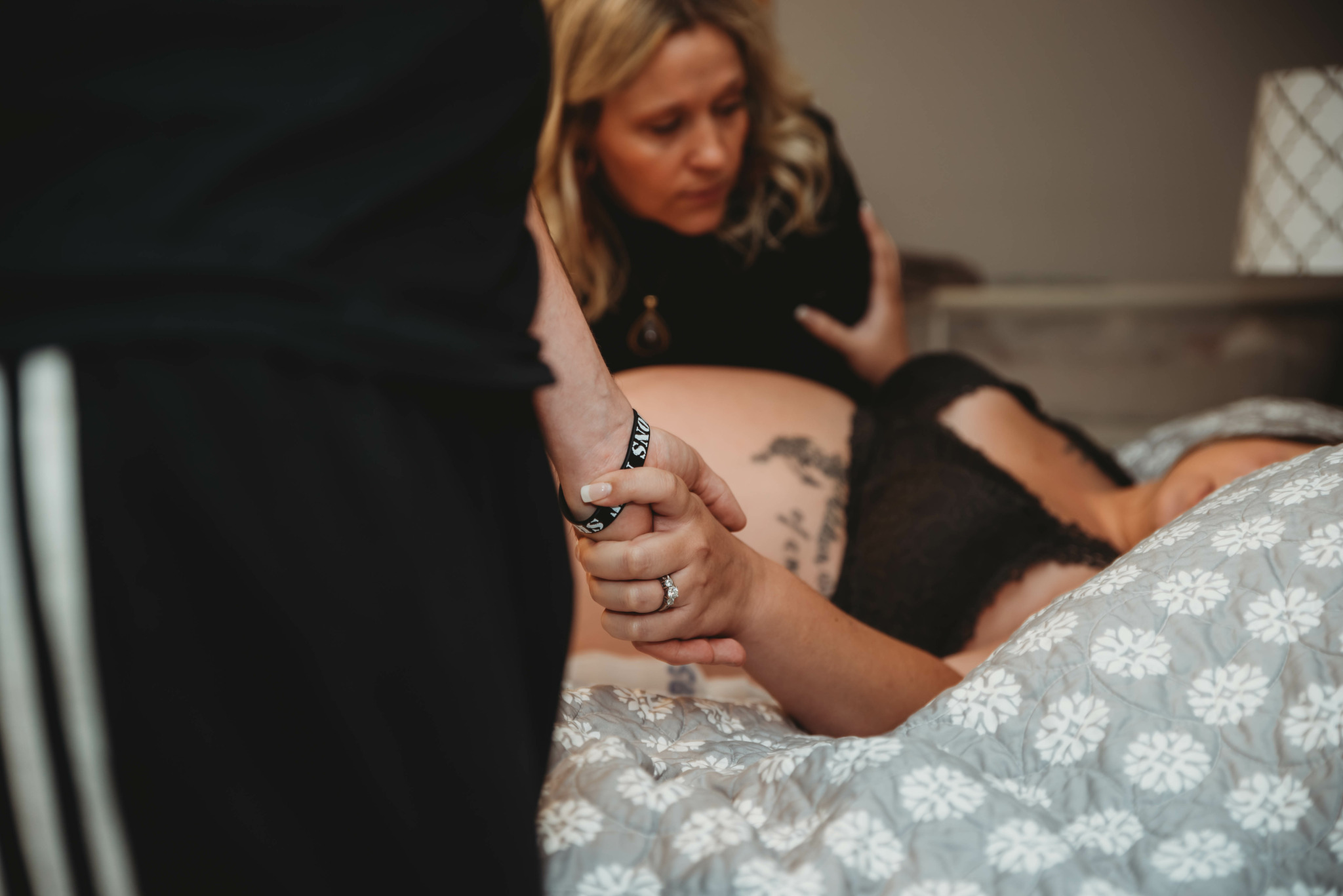 Woman with blonde hair looking at a person lying in bed, holding their hand, with a tattoo visible on their arm.