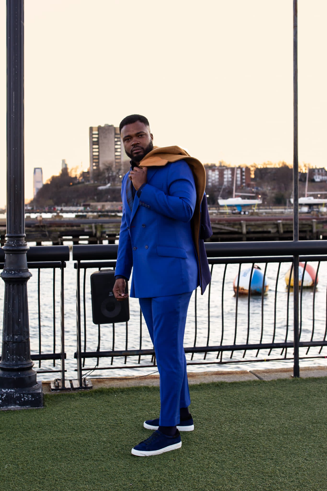 Man in blue suit standing near a railing by a river with city buildings in the background.