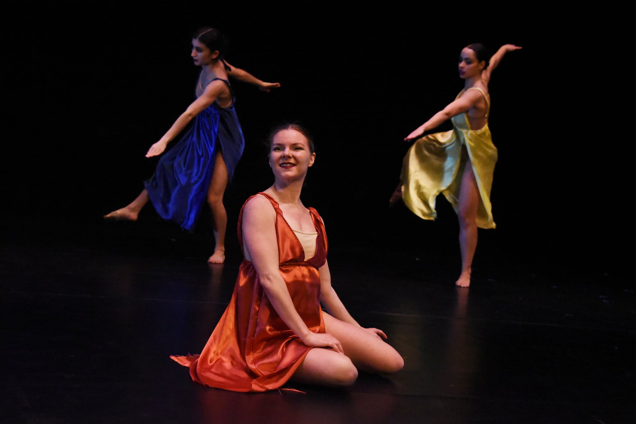 Three dancers on stage, one sitting in orange dress, two standing in blue and yellow dresses, performing ballet.