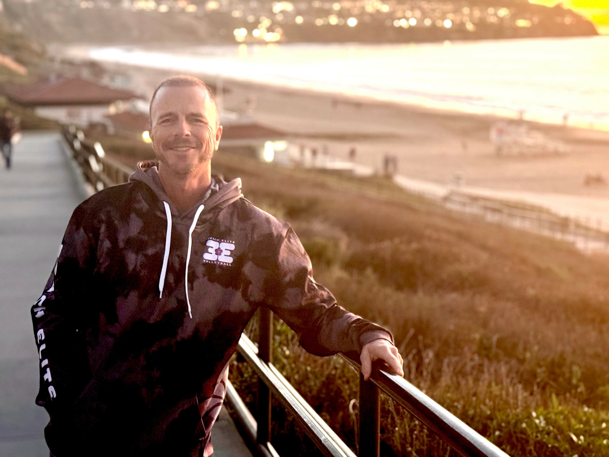 Man with beard smiling outdoors near a railing with a beach and sunset in background.