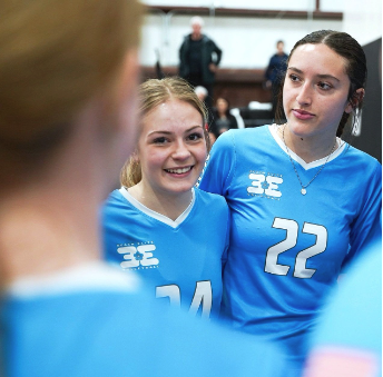 Two young women in blue sports jerseys with numbers 14 and 22, smiling and talking indoors.