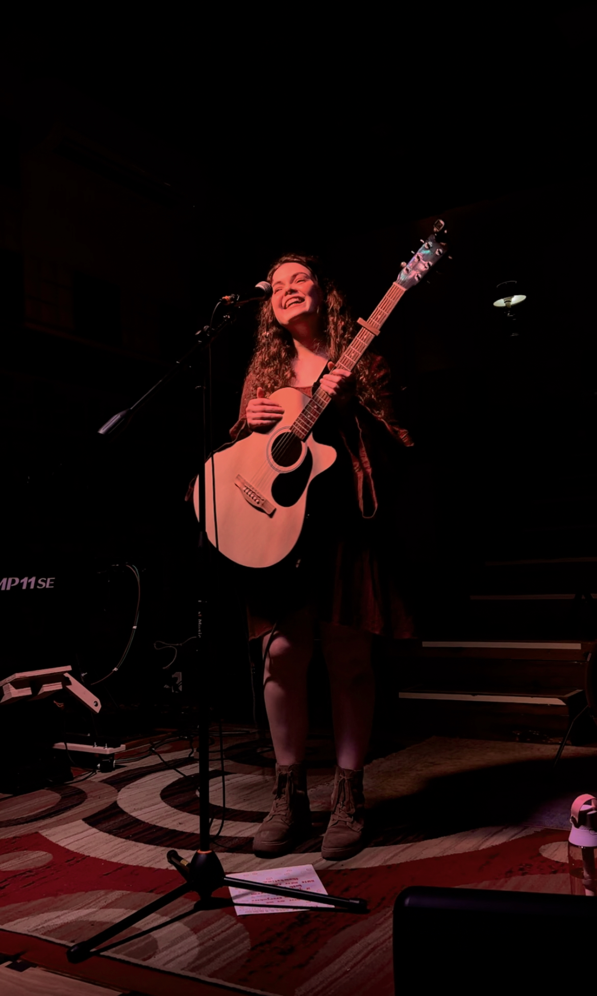 Person standing on stage holding an acoustic guitar, smiling, with a microphone and stage lights.