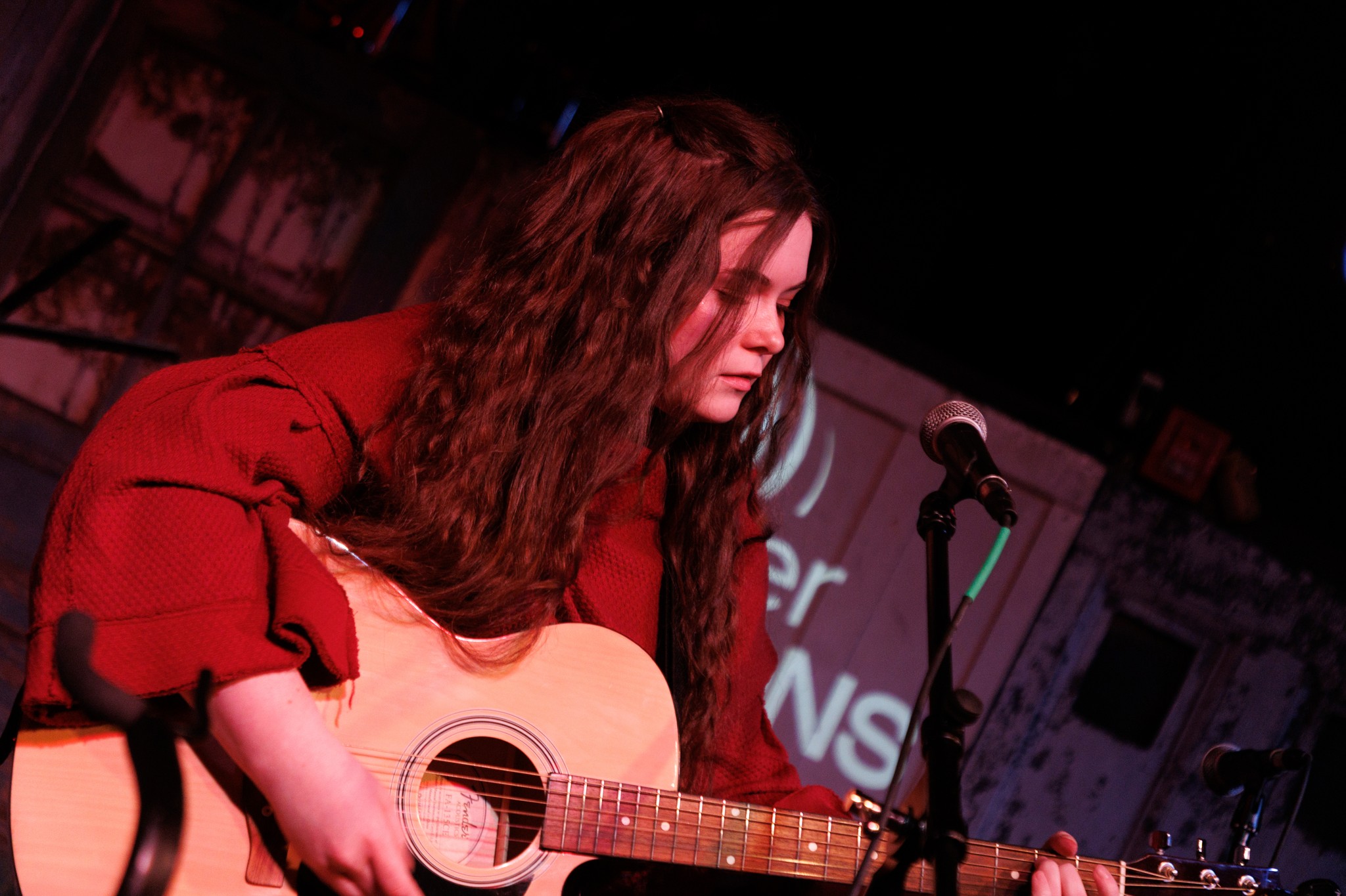Woman with long hair playing an acoustic guitar on stage with microphone nearby.