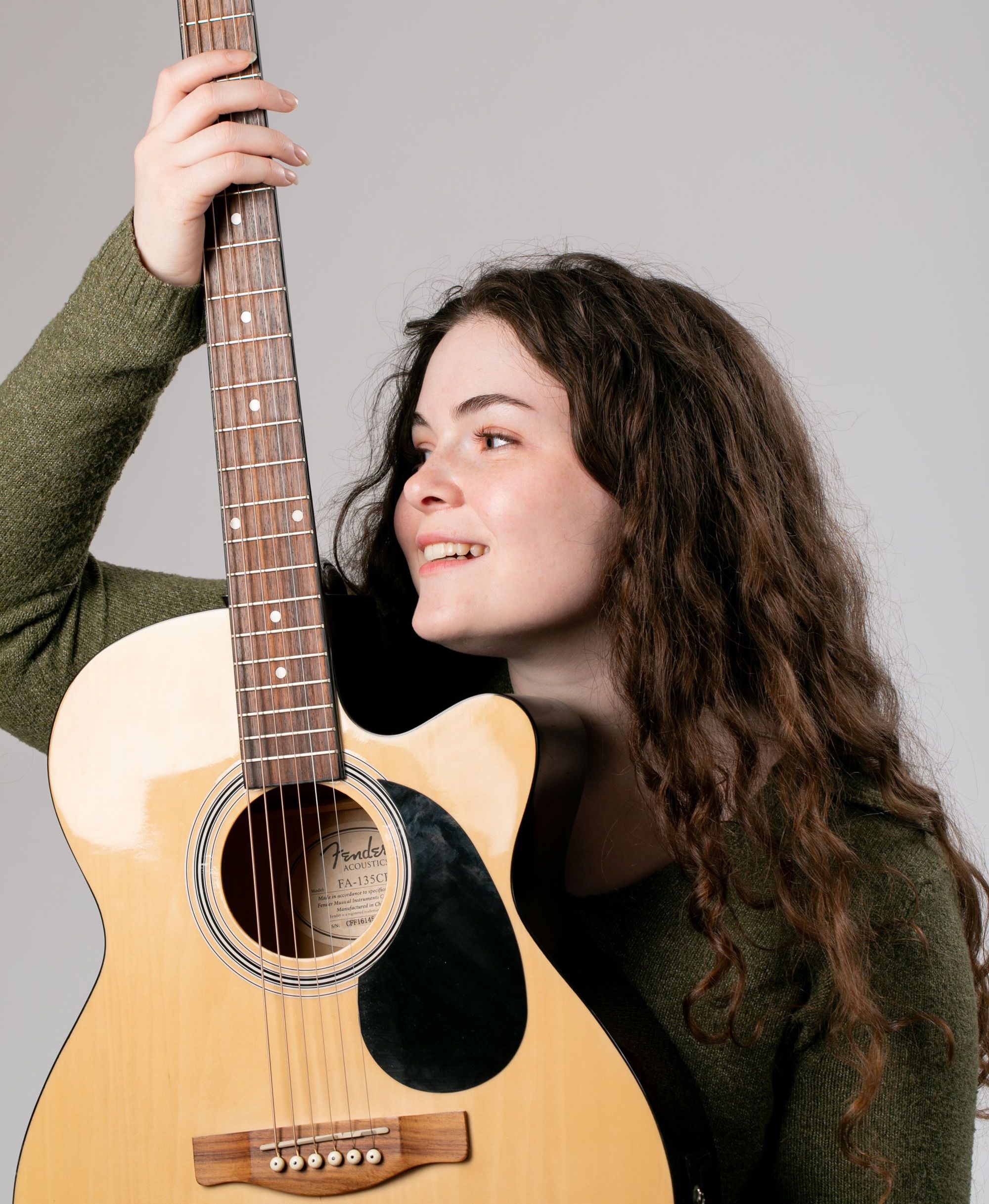Young woman with curly hair holding an acoustic guitar, smiling and looking to the side.