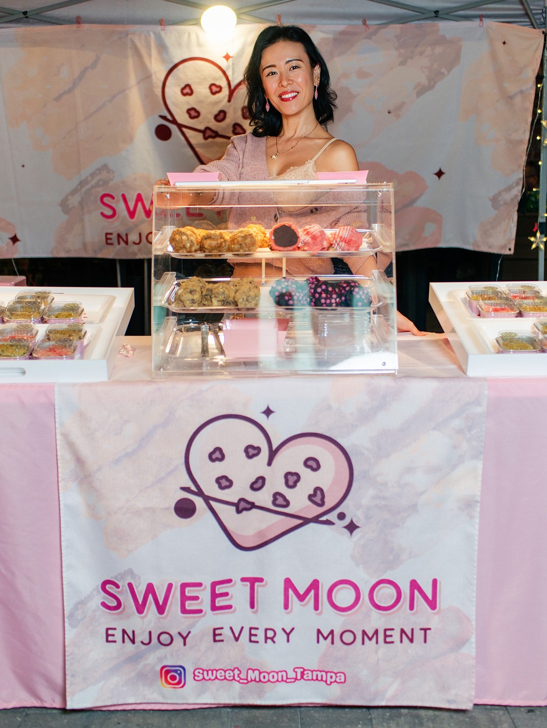 Woman smiling behind a clear display case with baked goods at a booth, pink and purple logo on tablecloth, banner in background.