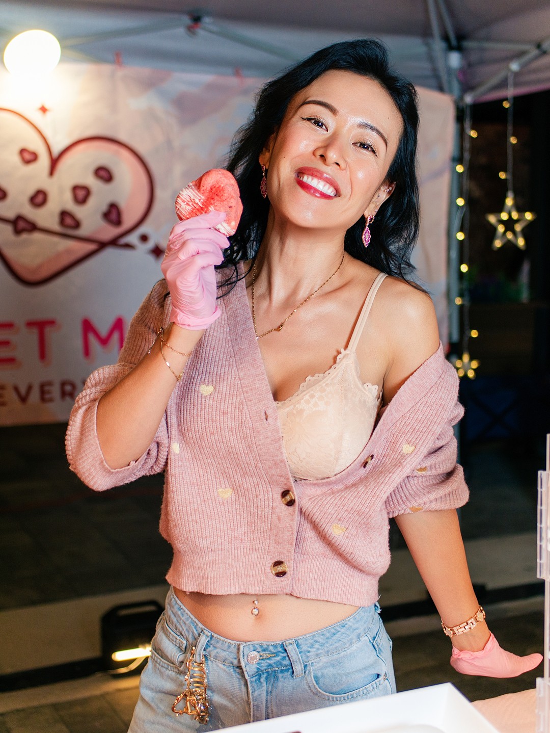 Smiling woman holding a pink heart-shaped object, wearing a pink cardigan and beige top, at festive event.