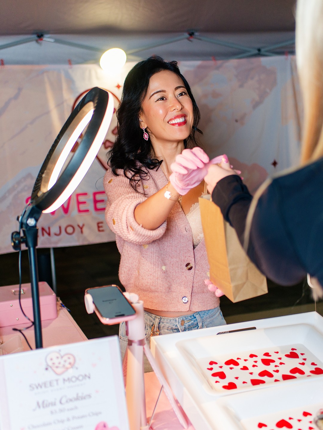 Young woman smiling at a booth, receiving a gift bag from a person, with a ring light and decorated backdrop.