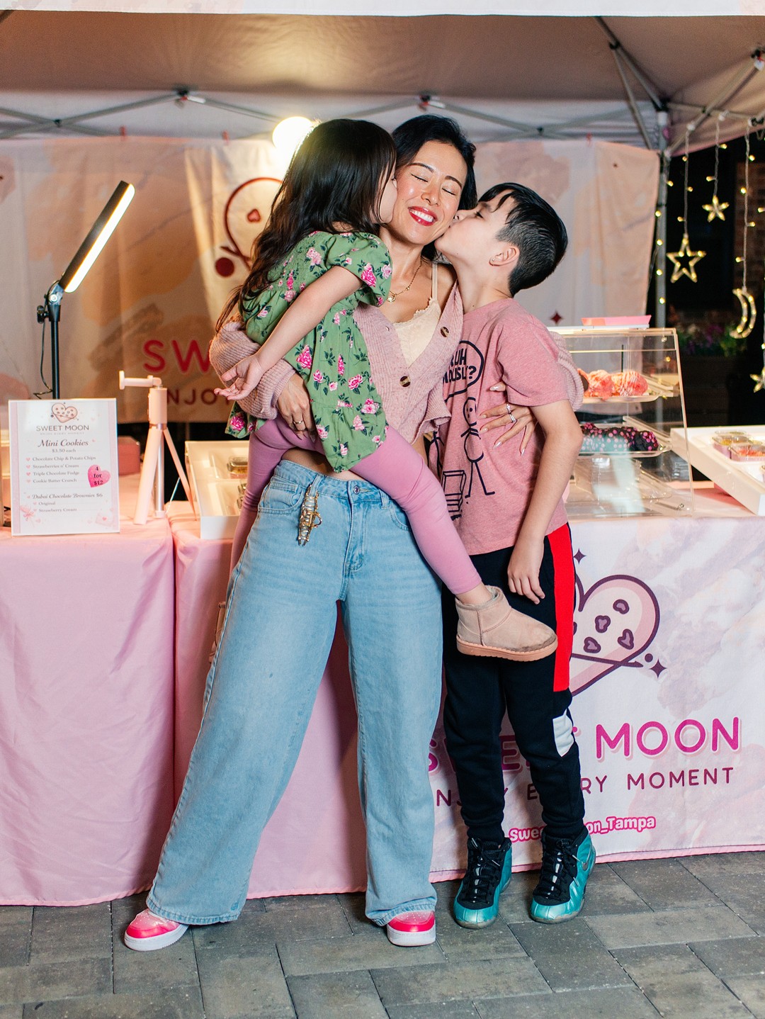 Woman with two children at a booth, one child hugging her, the other standing close, all smiling.