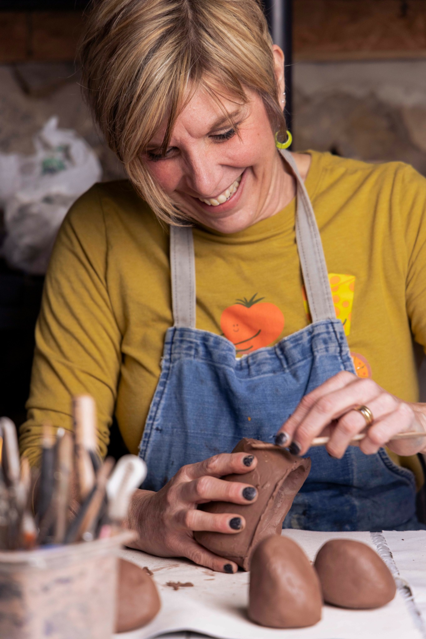 Woman smiling, wearing a yellow shirt and apron, working with clay in a pottery studio.