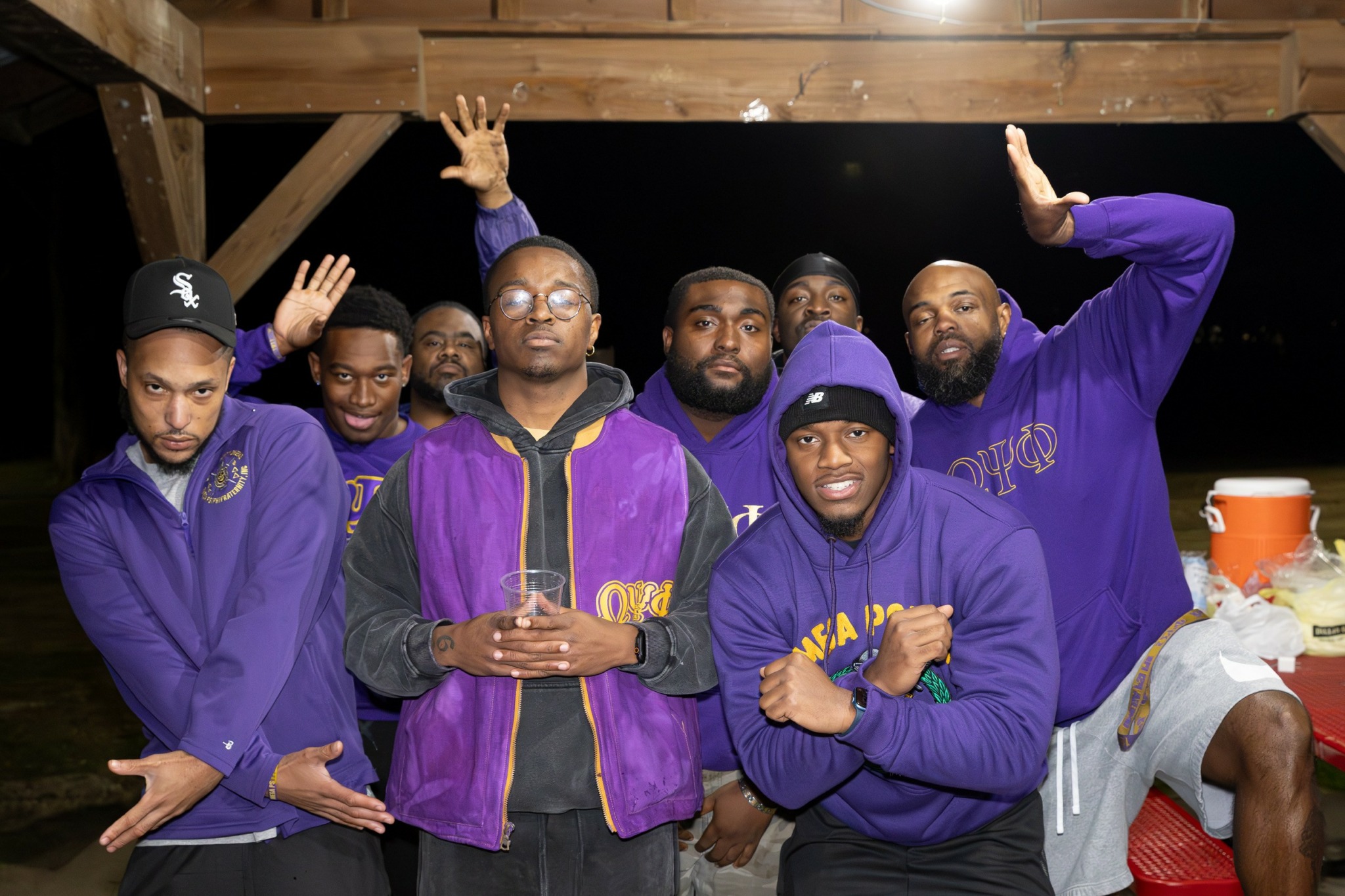 Group of seven men wearing purple and black jackets, some raising hands, in a wooden outdoor setting at night.