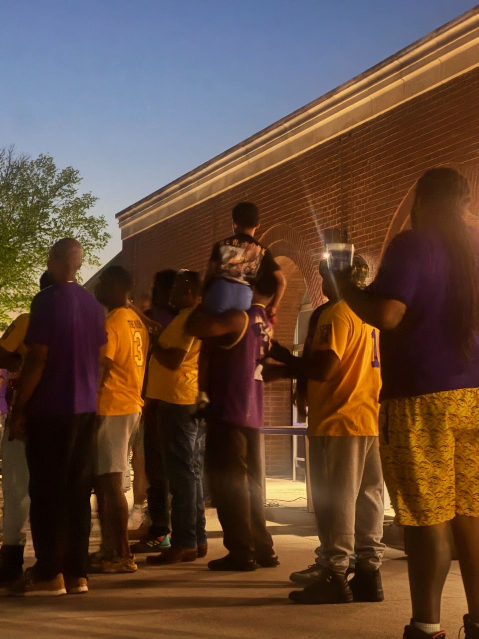Group of people gathered outdoors in front of a building, some wearing yellow shirts, one person carrying a child on their shoulders.