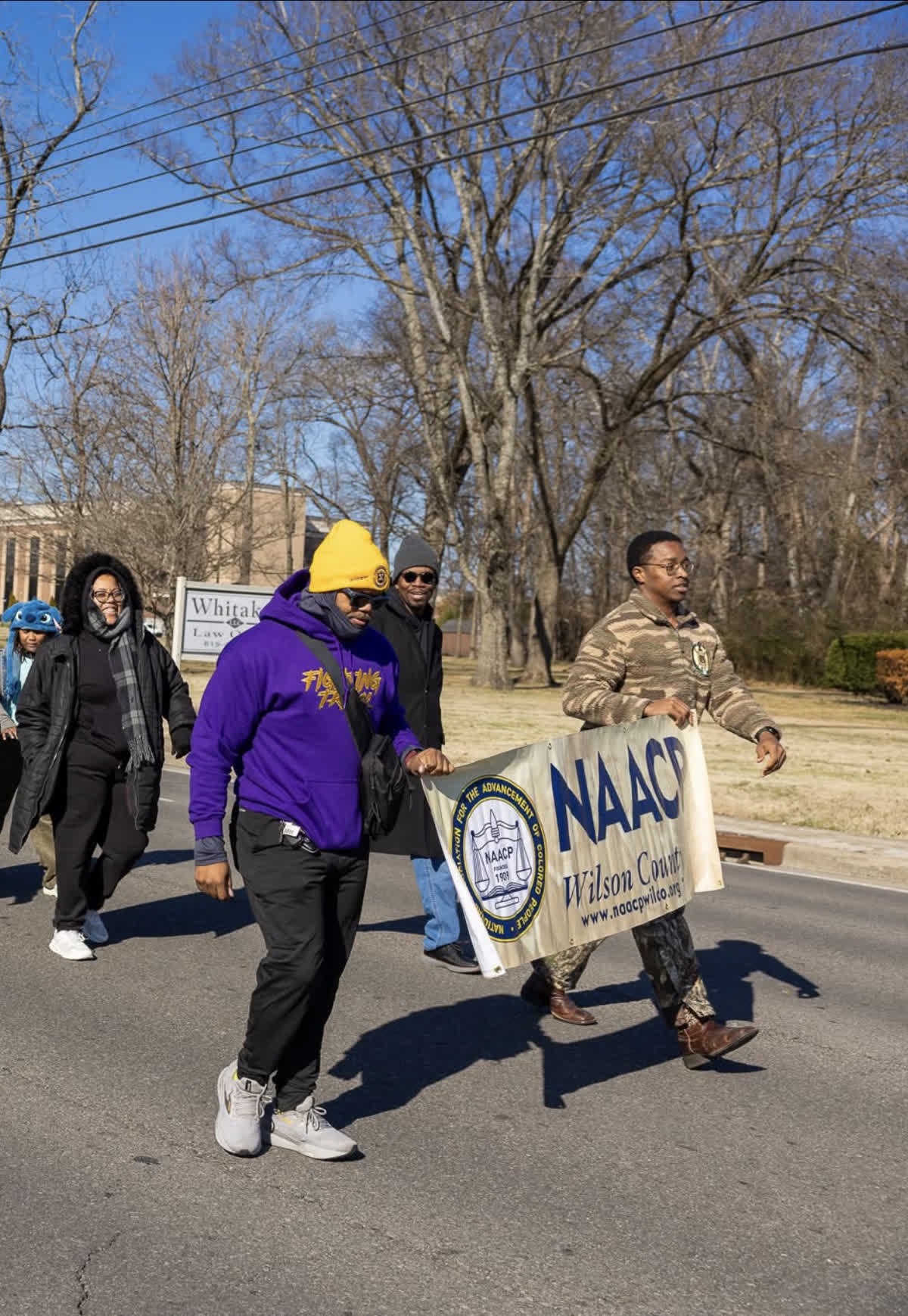 Group of people walking on a street holding a NAACP banner, trees and power lines in background.