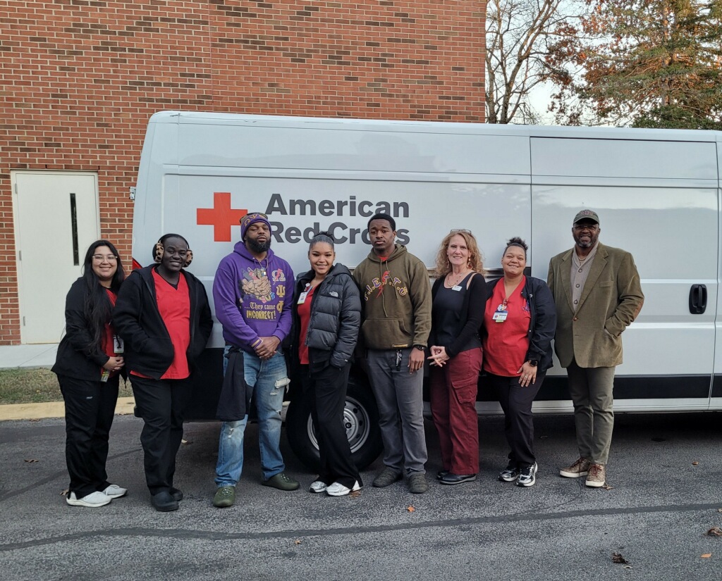Group of eight people standing in front of a Red Cross vehicle with a brick building in background.