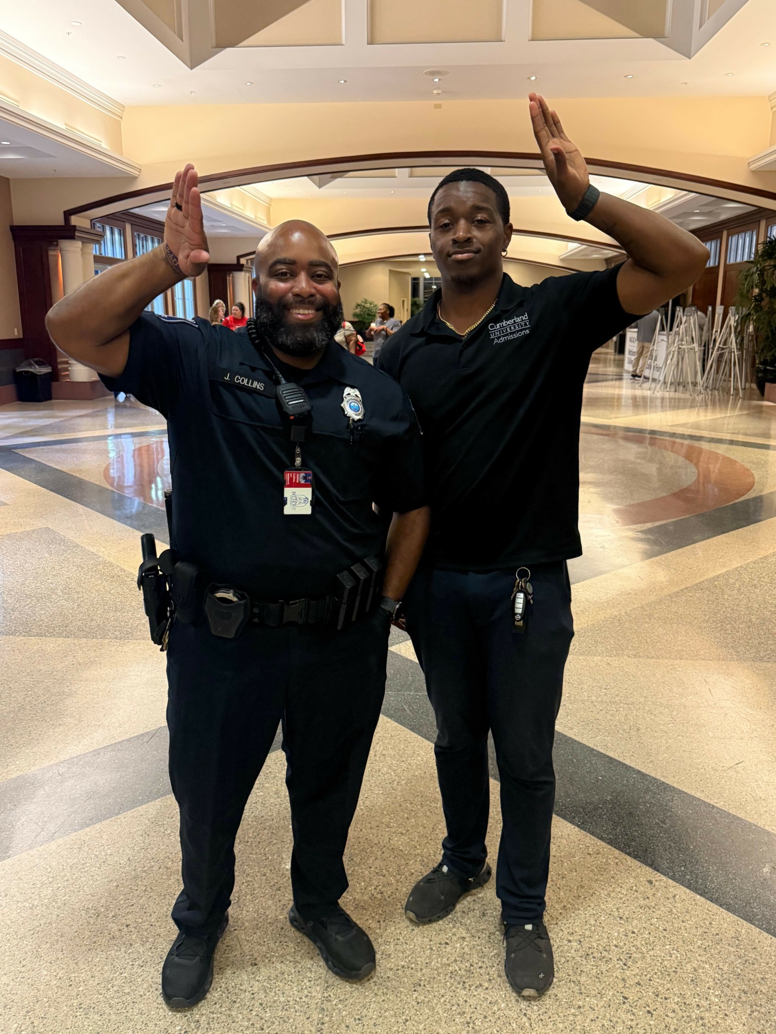 Two men in uniform standing inside a building, raising their hands in a celebratory gesture, smiling at the camera.