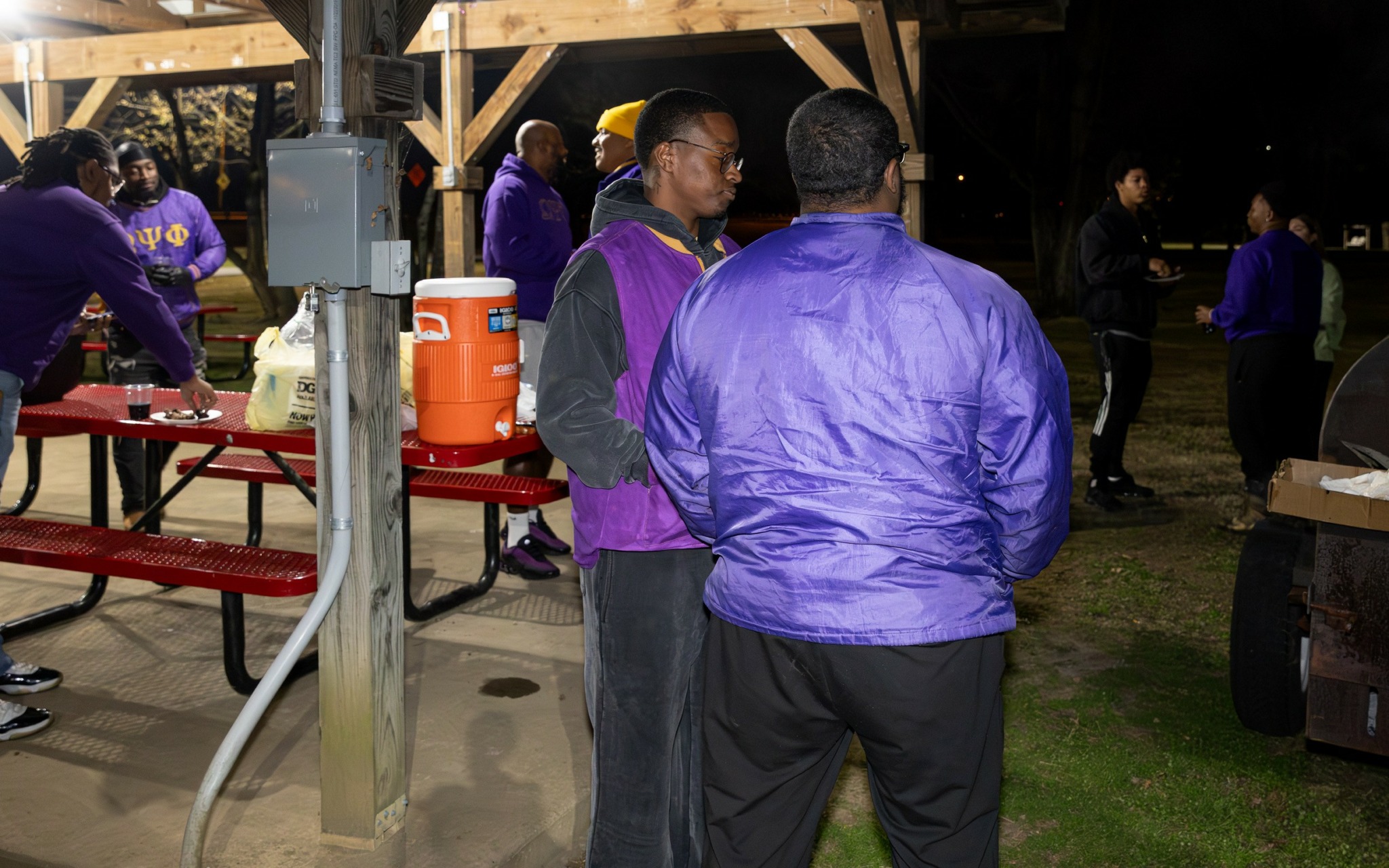 Group of people gathered outdoors at night, some wearing purple jackets, near a table with food and drinks.