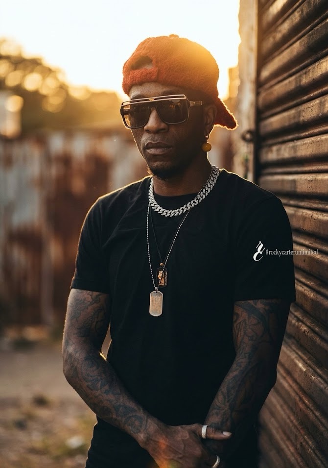 Young man with orange bandana, sunglasses, black t-shirt, and jewelry stands outdoors near a wooden wall.
