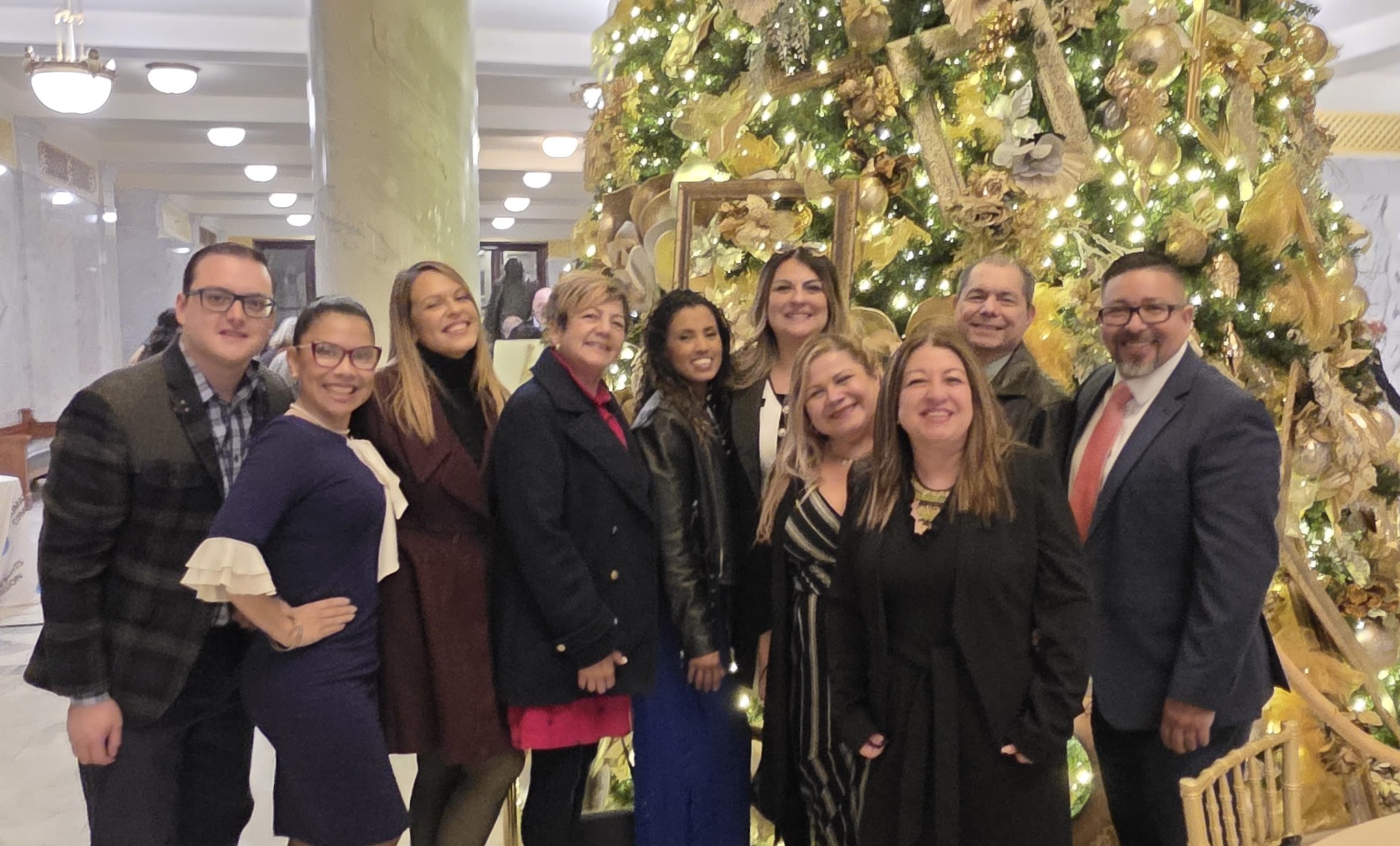 Group of ten people smiling in front of a decorated Christmas tree indoors.