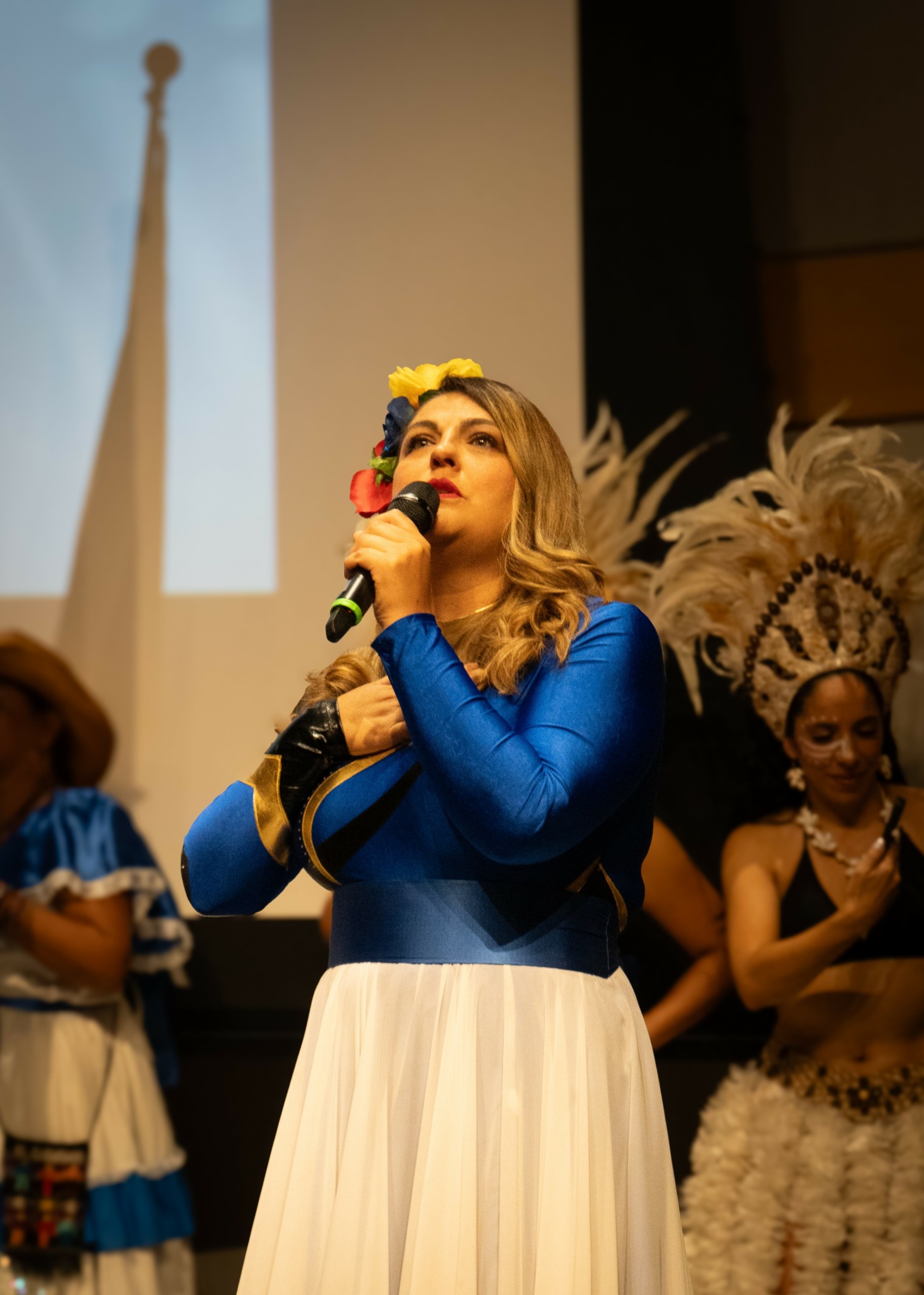 Woman in blue and white dress with floral headpiece holding a microphone on stage, with other performers in background.