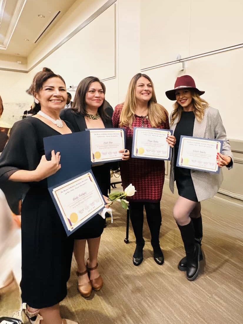 Four women standing indoors, holding certificates, smiling, and posing for a photo.