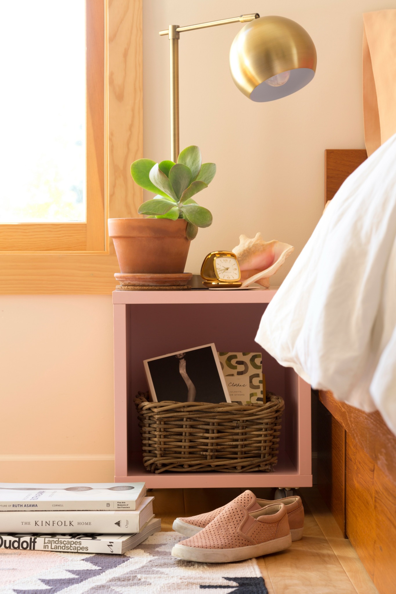 Nightstand with a potted plant, gold desk lamp, and a small clock next to a bed with white bedding.