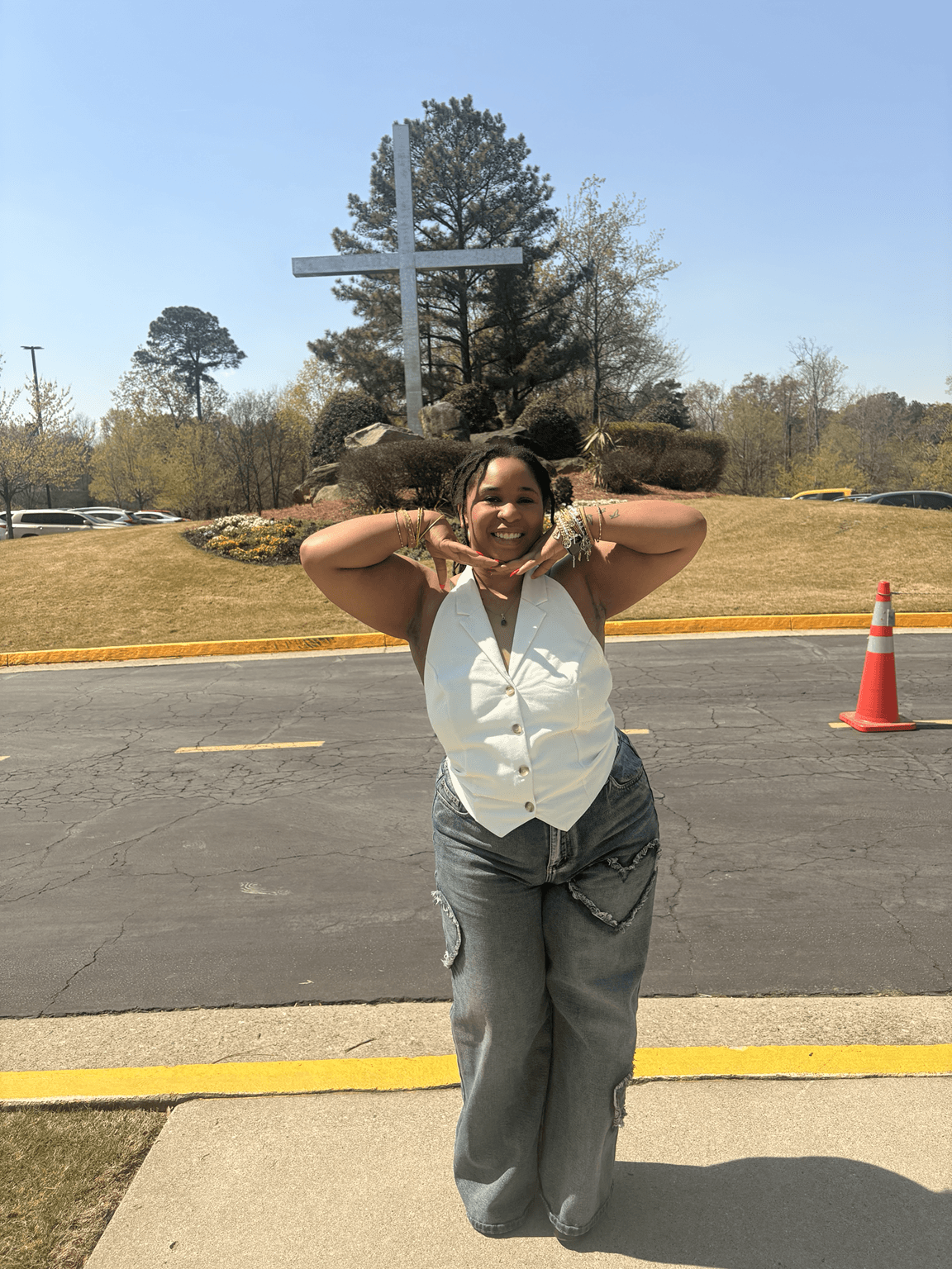 Woman standing outdoors on sidewalk, smiling, making peace signs with both hands, trees and a cross monument in background.