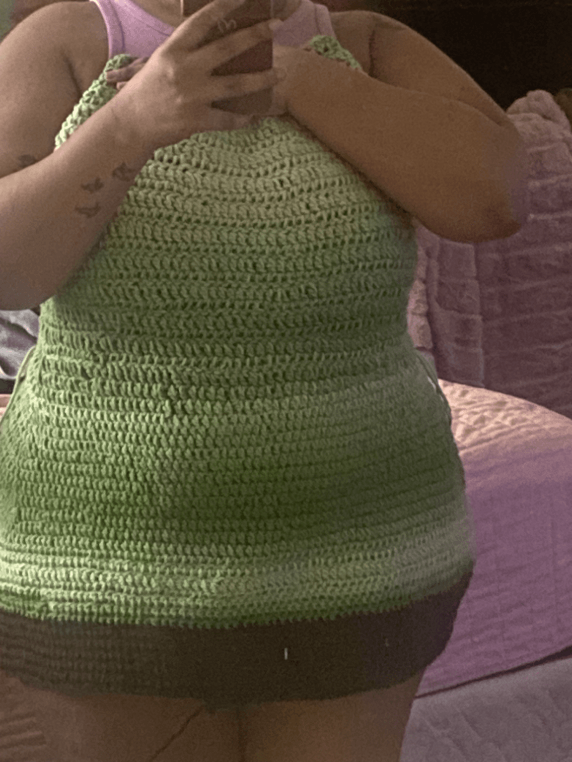 Person taking a mirror selfie wearing a green and black crocheted dress, holding phone with left hand, arm tattoos visible.