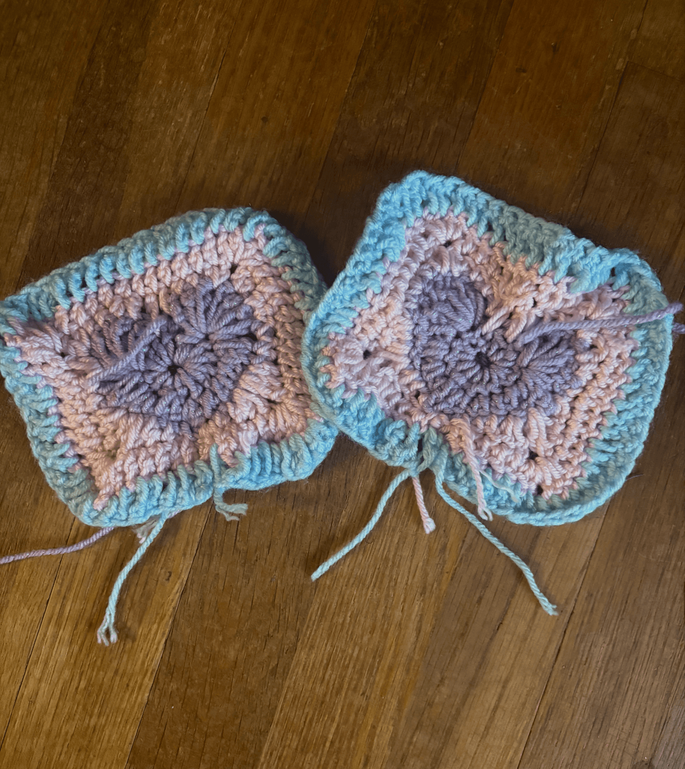 Two crocheted square pieces with pink and purple yarn, bordered by blue yarn, on a wooden surface.