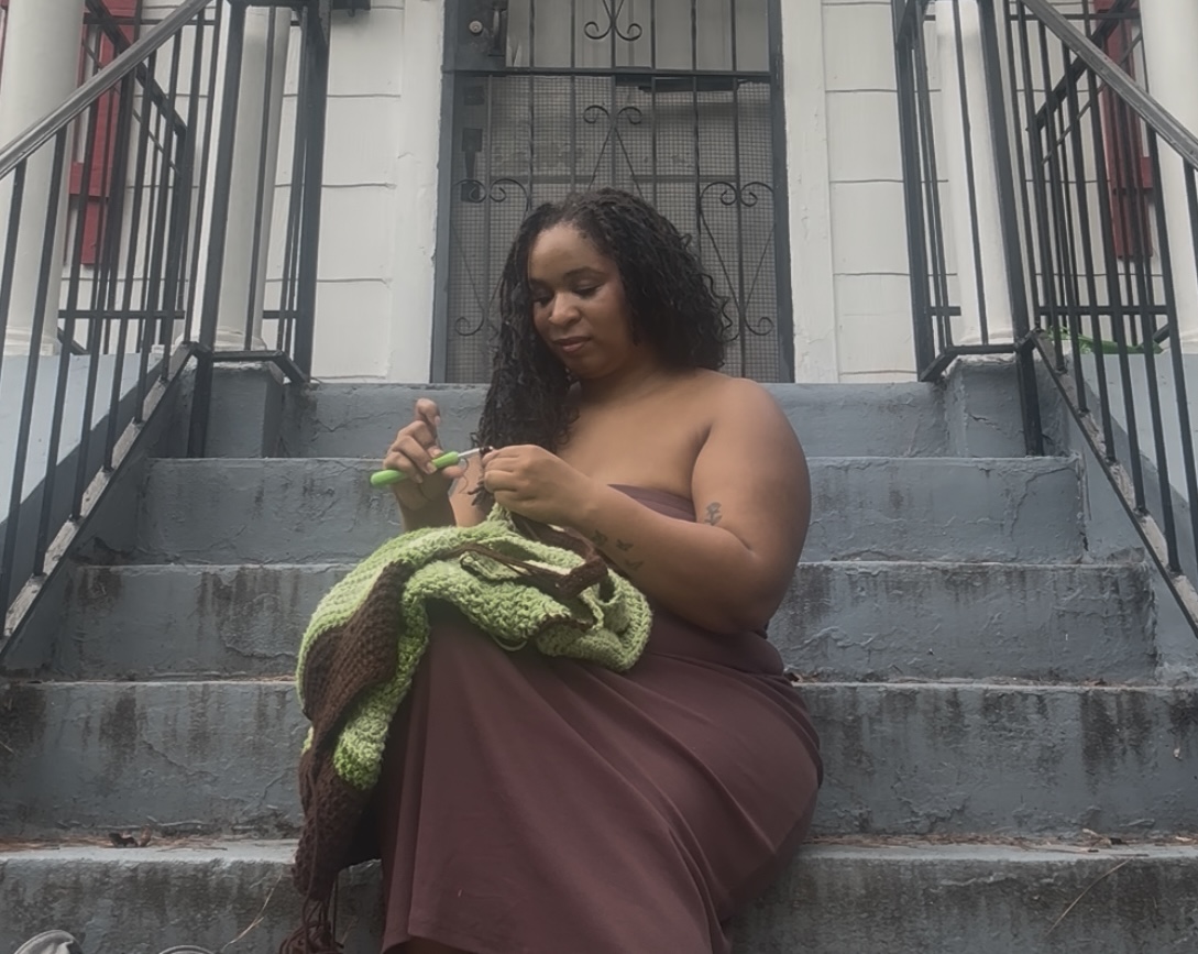 Woman sitting on stairs, knitting with a green and brown blanket, in front of a building with a door and railing.
