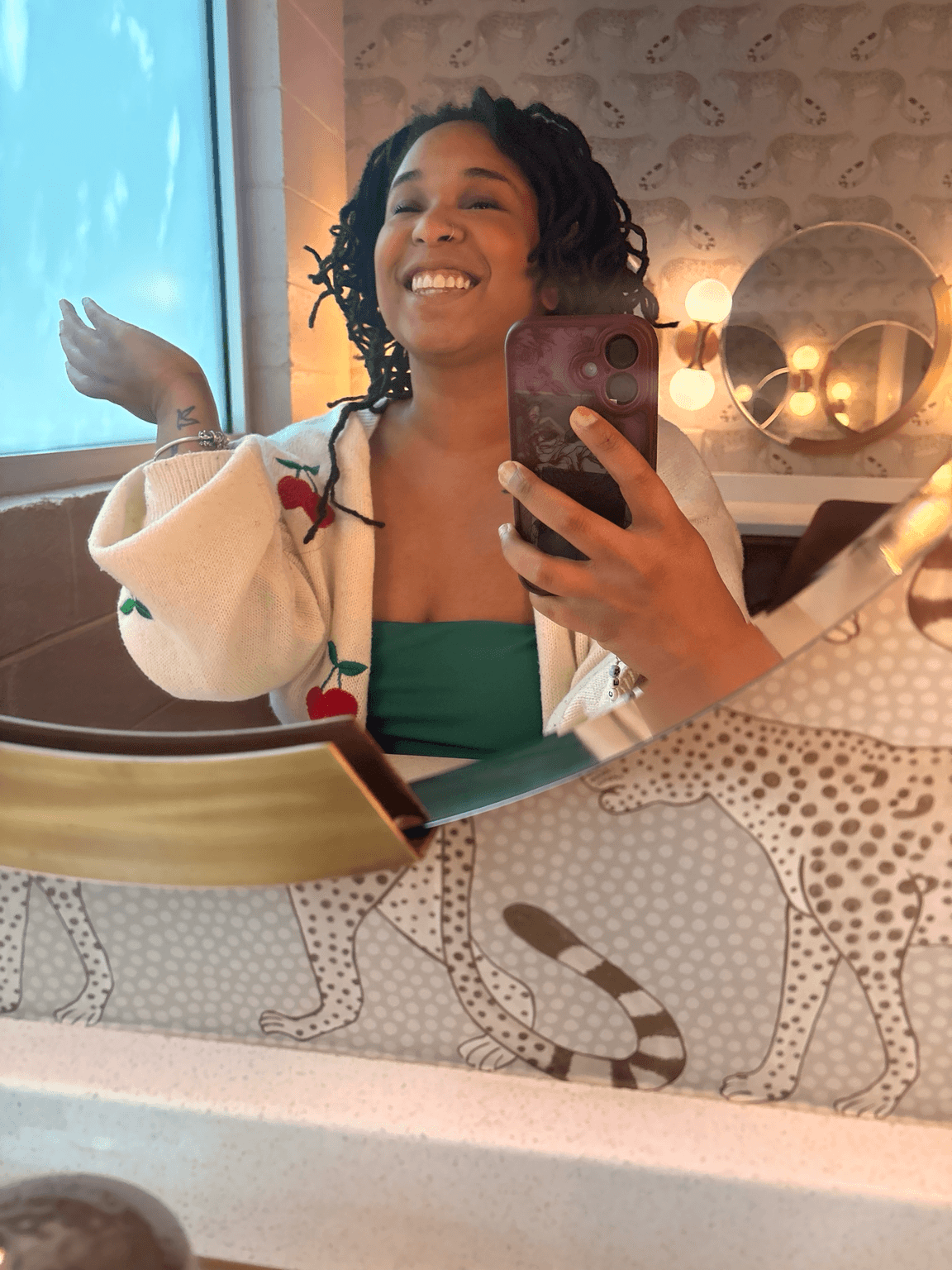 Woman smiling in mirror, holding phone, wearing green top and cream cardigan, in room with decorative wall and mirror.