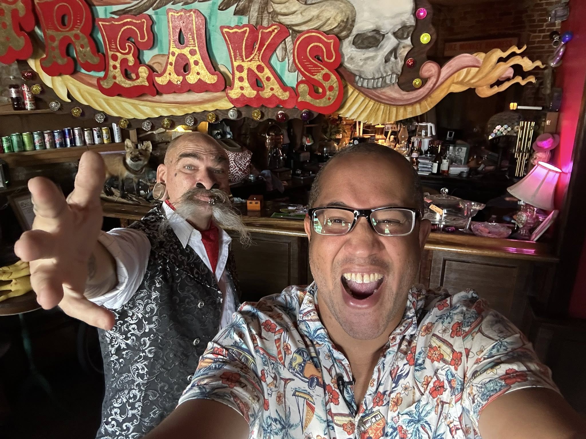 Two men smiling and taking a selfie in a bar with colorful decorations and a skull in the background.