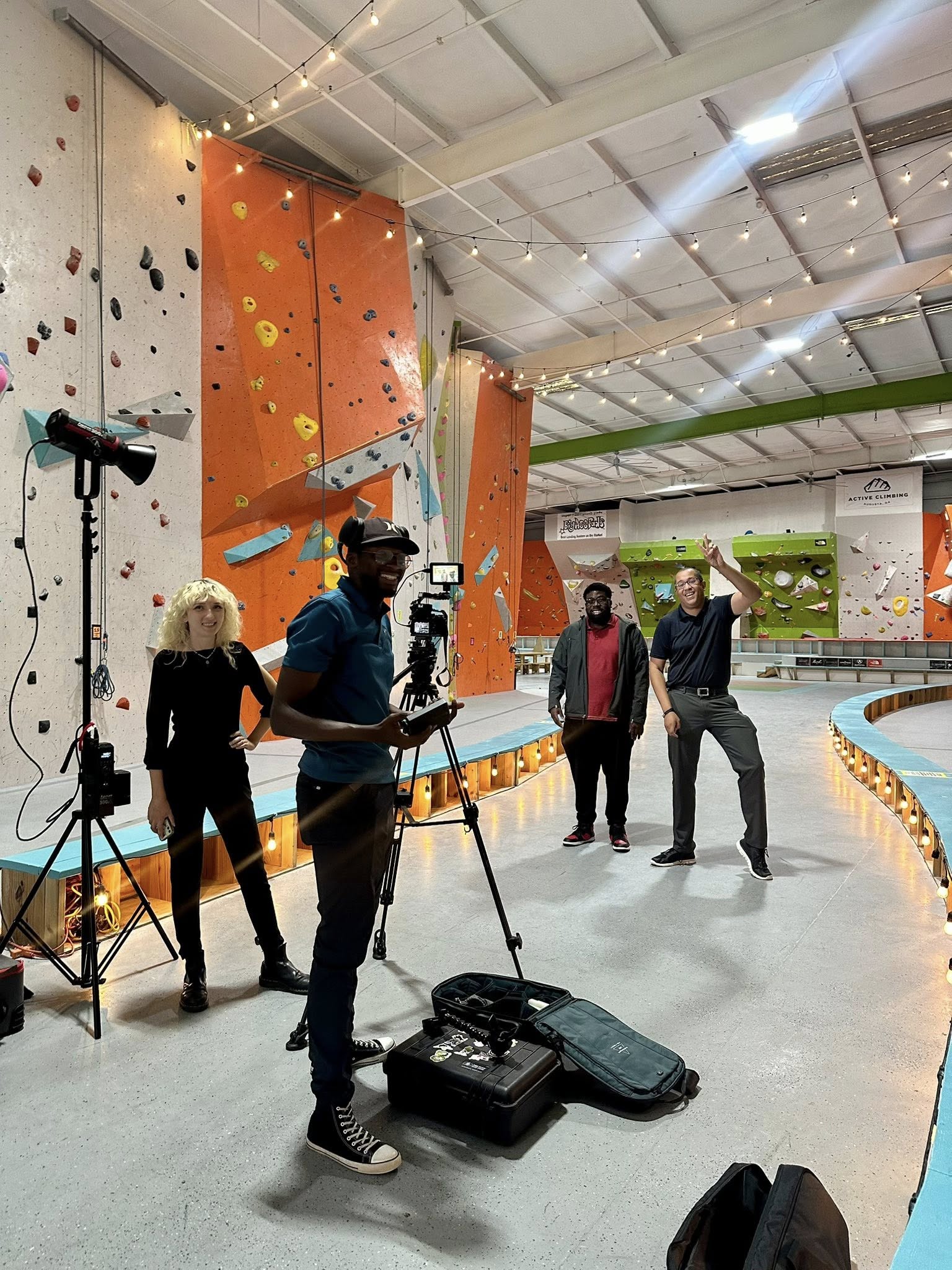 Indoor climbing gym with four people, one person filming, and climbing walls in background.