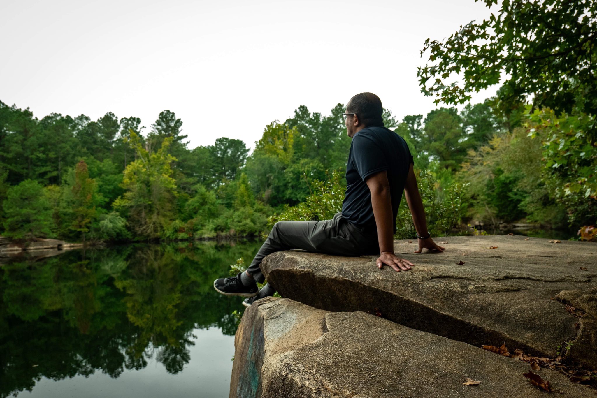 Person sitting on a large rock by a calm river, surrounded by green trees, looking at the water.