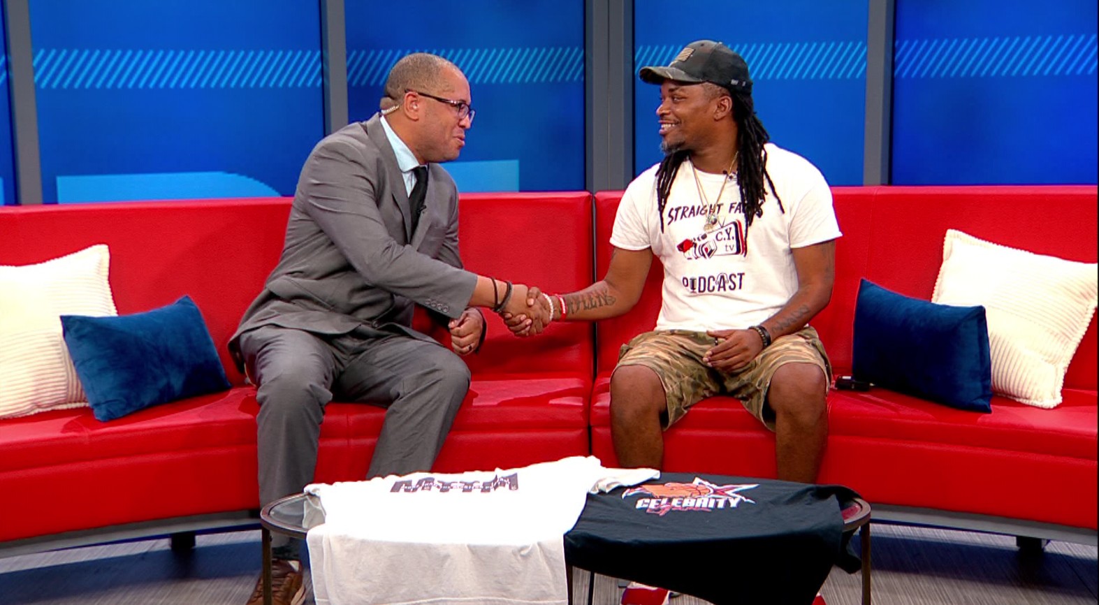 Two men shaking hands on a red couch in a studio setting with blue background and pillows.