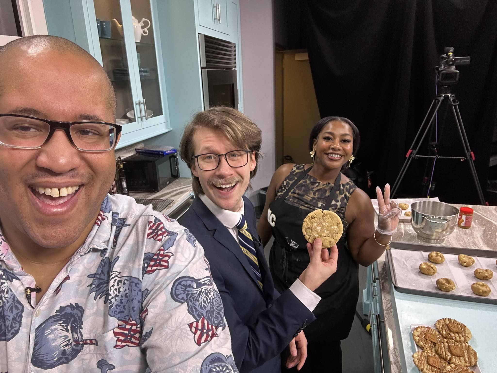 Three people smiling in a kitchen, one holding a cookie, with baking trays and camera equipment nearby.