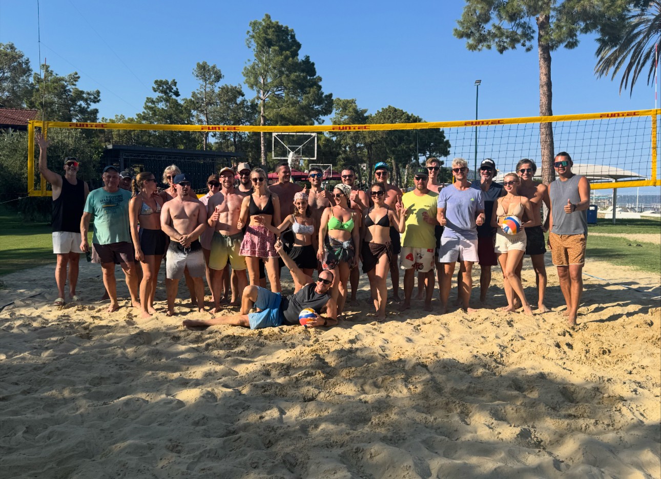 Group of people on a sandy volleyball court outdoors, some holding volleyballs, with trees and a blue sky in background.