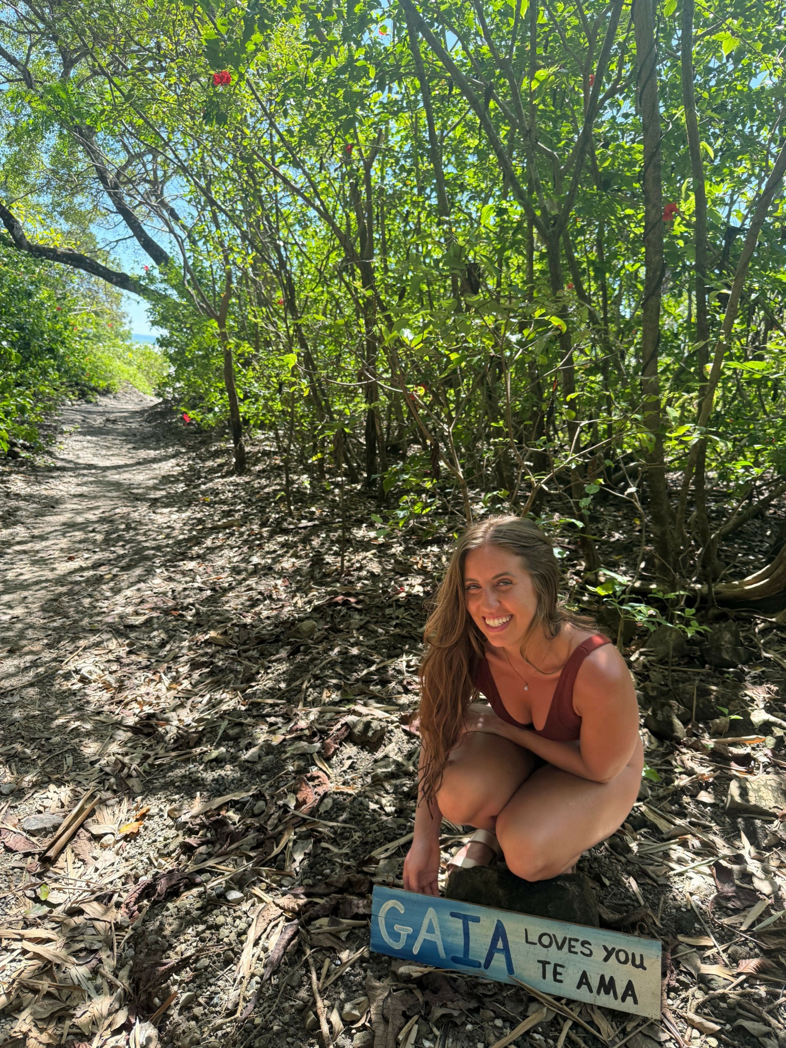Young woman crouching on a dirt path surrounded by green trees and foliage, smiling at the camera, holding a sign.