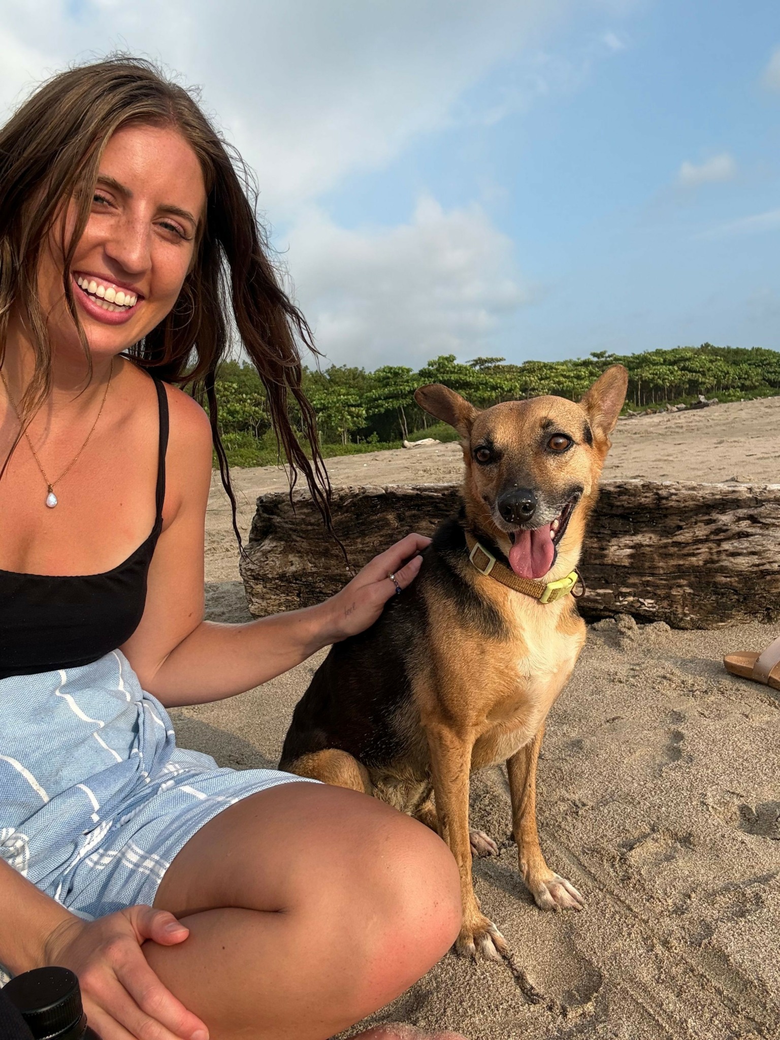 Woman with long hair smiling next to a dog on a sandy beach with trees and sky in background.