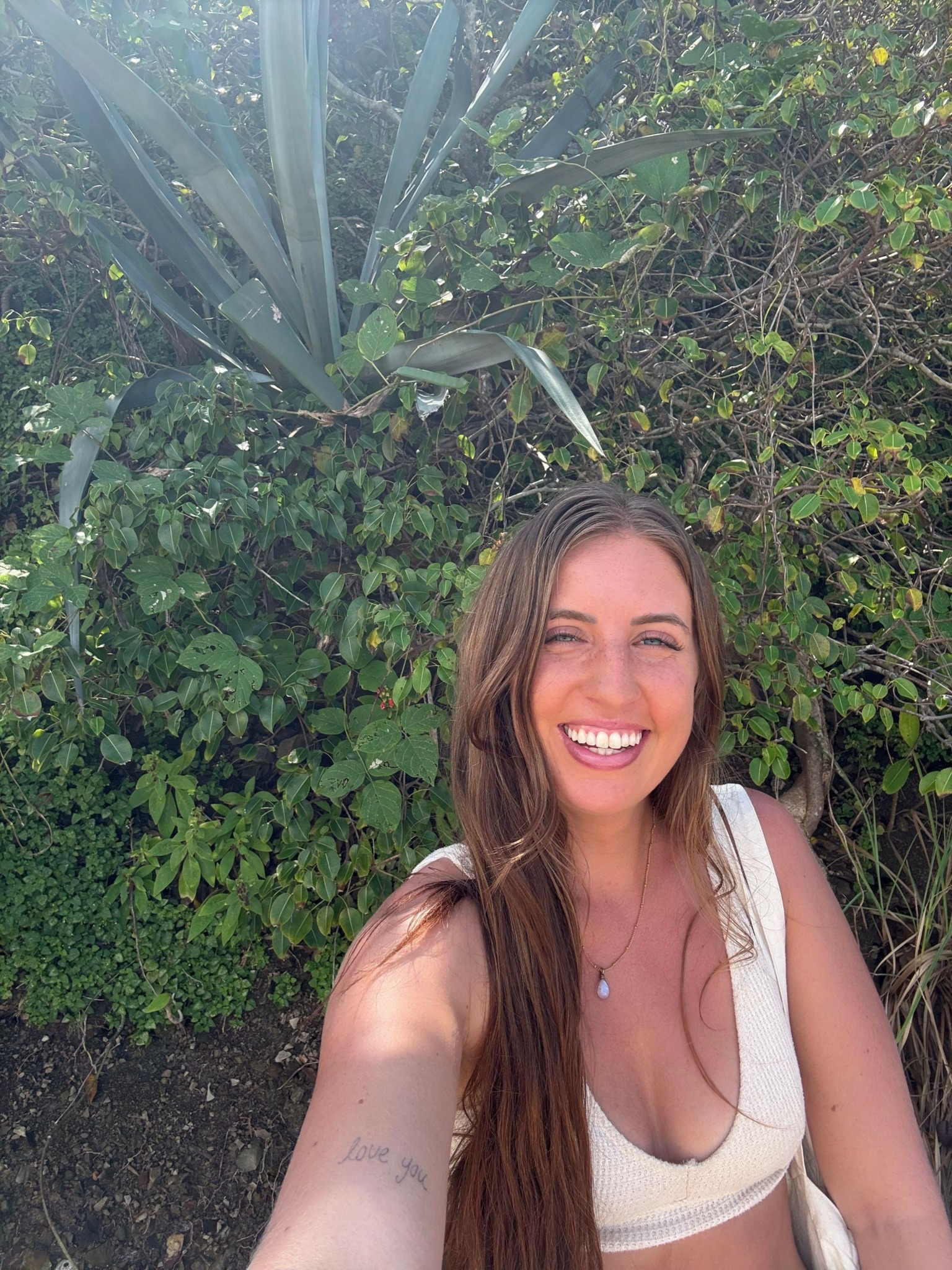 Young woman with long hair smiling outdoors in front of green foliage.