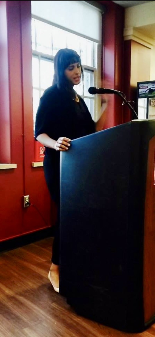 Woman with dark hair and bangs speaking at a podium in a room with red walls and large window.