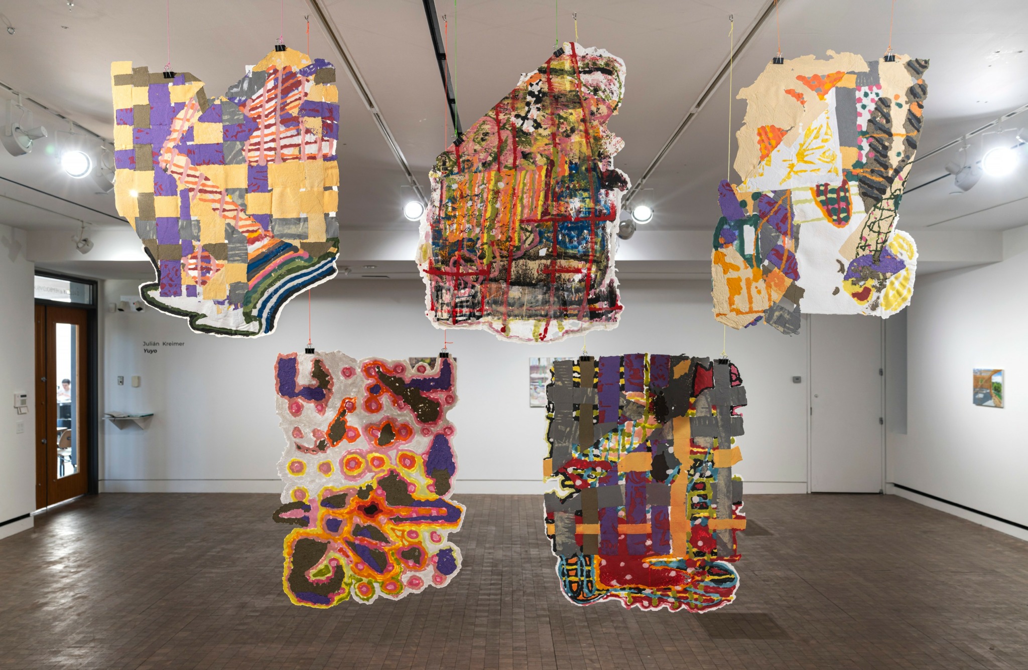 Six colorful abstract textile art pieces hanging from the ceiling in a gallery room with white walls and wooden floor.