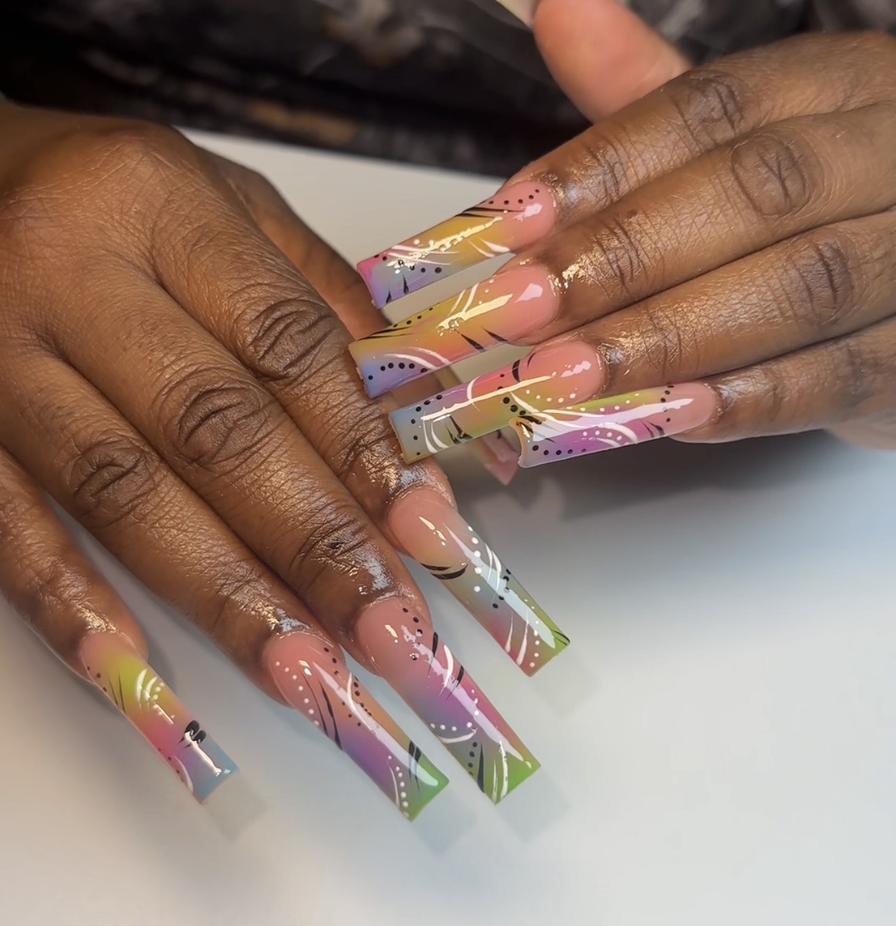 Close-up of hands with colorful, decorated long acrylic nails resting on a white surface.